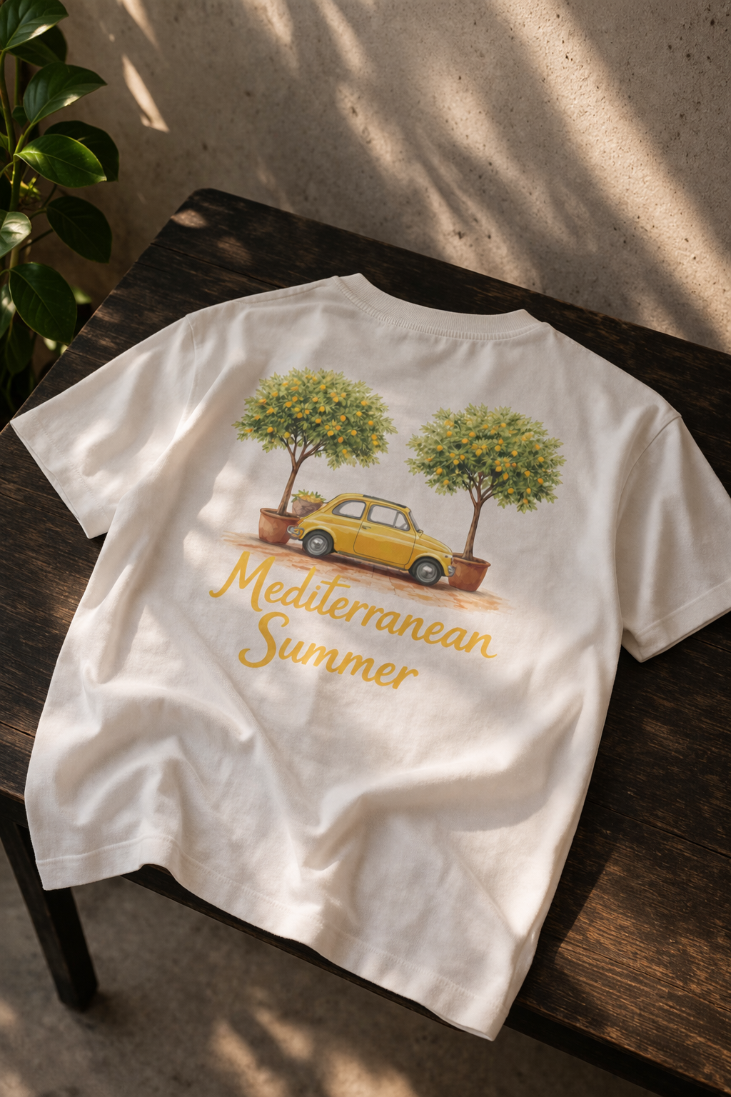 Wit t-shirt met een losse pasvorm. op de rug van het t-shirt staat een ontwerp met een klassieke gele fiat 500 met 2 mooie grote citroenbomen op een terracotta ondergrond. onder het ontwerp staat in sierlijke gele letters Mediterranean Summer. presentatie op houten tafel met betonnen muur.