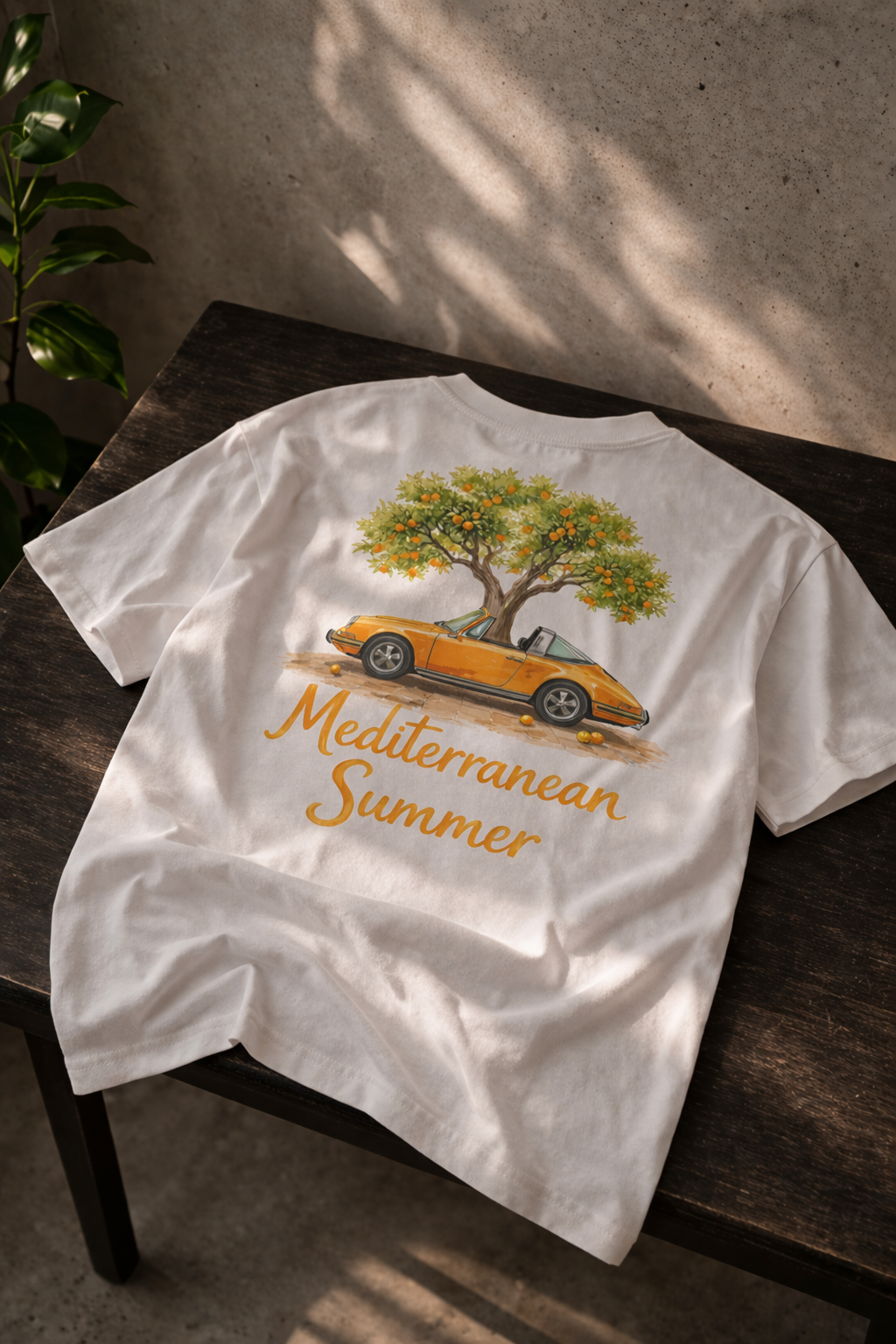 Wit t-shirt met een losse pasvorm. op de rug staat een design van een oranje klassieke Porsche 964 targa met een grote mooie sinaasappelboom. onder het ontwerp staat in sierlijke letters Mediterranen Summer. presentatie op een mooie houten tafel met betonnen muur. 